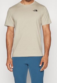 Muž v béžovém tričku s krátkým rukávem s černým logem "The North Face" a modrých sportovních kalhotách s logem La Sportiva na stehně.