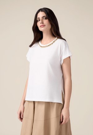 Top corto bianco a maniche corte con scollatura accentuata in oro, spacchi laterali e una vestibilità rilassata, abbinato a una gonna beige fluida.