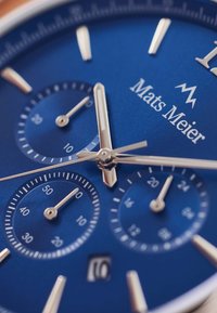 Blaues Zifferblatt-Chronographen-Armbanduhr mit silbernen Zeigern und Ziffernblättern, ausgestattet mit einem Datumsdisplay, dem Markennamen "Mats Meier" und strukturiertem Hintergrund.