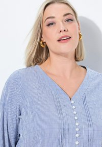 Modrá a bílá pruhovaná blůza s výstřihem do V, zapínáním na knoflíky a texturovanou tkaninou. Výrazné doplňky zahrnují zlaté kruhové náušnice.