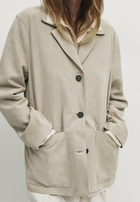 Beige mokkanahkatakki, jossa on kaulus, kolme mustaa nappia, kaksi etutaskua ja rento malli. Pehmeä rakenne ja rento muotoilu.