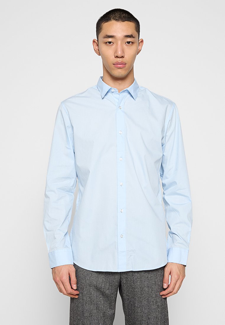 Giovane uomo vestito con una camicia azzurro chiaro e pantaloni grigi, in piedi davanti a uno sfondo grigio chiaro uniforme.