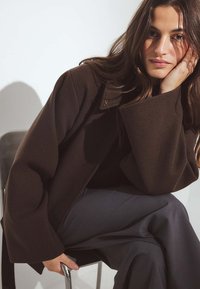 Hnedá prešívaná bunda z vlnenej zmesi s klopovým golierom, skombinovaná s tmavošedými voľne strihanými nohavicami, ktoré ukazujú hladké textúry a uvoľnený štýl.