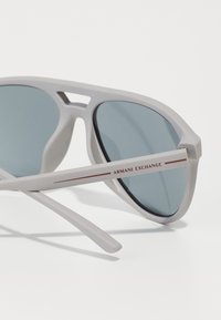 Valged plastist pilooti stiilis prillid tumedate läätsedega. Raamil on iseloomulik lõike ning punane "Armani Exchange" brändimine.