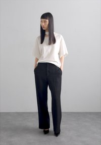 Bela oversize majica s kratkimi rokavi, v kombinaciji s črnimi krojenimi hlačami. Model stoji samozavestno ob nevtralni ozadju.