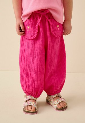 Dítě nosí jasně růžové texturované kalhoty s předními kapsami a květované sandály se dvěma sponami.