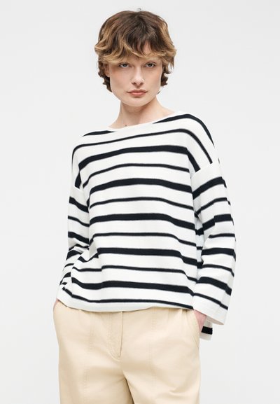 Άτομο με κοντά κυματιστά μαλλιά, φορώντας μακρυμάνικη μπλούζα με μαύρες και άσπρες οριζόντιες ρίγες και μπεζ παντελόνι, με τα χέρια στις τσέπες.