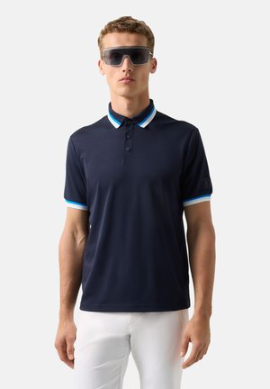 Młody mężczyzna w dużych, ciemnych okularach przeciwsłonecznych, granatowej koszulce polo z niebiesko-białym paskowanym kołnierzykiem i rękawami oraz białych spodniach.