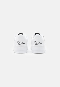 White sneakers with black accents, smooth leather upper, flat sole, and a signature design detail on the heel. Minimalist and structured shape.