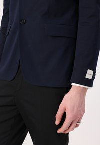 Marineblazer met één zwarte knoop, twee voorzakken en witte manchetten zichtbaar. Kleine zilveren Calvin Klein-tag op de mouw. Zwarte broek.