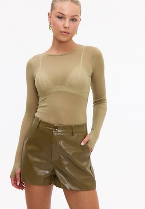 Femme portant un top à manches longues translucide couleur olive montrant un soutien-gorge en dentelle en dessous et un short en cuir vert foncé taille haute, la main dans la poche.