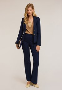 Marineblauw op maat gemaakt pak met gouden knopen, beige V-hals top en een statement ketting. Model houdt een clutch en draagt lichtgekleurde schoenen.