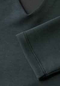 Tela verde oscuro con una textura suave, presenta un escote en V y un puño de manga doblado. Se pueden ver los detalles de costura a lo largo del borde.
