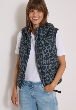 Femme souriante portant une veste matelassée imprimé léopard vert et noir, un t-shirt blanc et un jean bleu clair avec une main dans la poche.