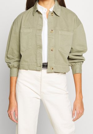 Mujer con chaqueta corta de color oliva claro con botones en la parte delantera, grandes bolsillos en el pecho, mangas fruncidas, camisa blanca, pantalones blancos de talle alto y cinturón con tachuelas.