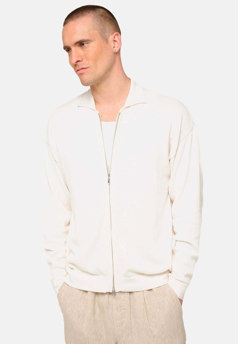 Pull zippé beige clair avec une texture côtelée, doté d'un col montant et d'une coupe décontractée, associé à un pantalon en lin beige.