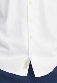 Chemise en lin blanc avec un devant boutonné, des boutons de couleur claire et une étiquette cousue en bas à gauche. Tissu texturé et respirant.