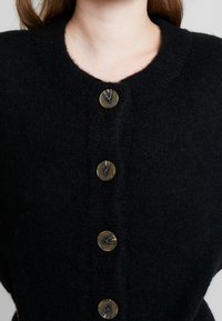 Svart stickad cardigan med rund halsringning och fem stora, texturerade knappar i ett kontrasterande material ned längs framsidan.