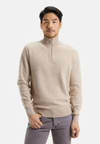 Sweater beige à col zippé avec un tricot texturé, des poignets et un ourlet côtelés, associé à un pantalon gris clair, arborant un logo minimaliste sur la poitrine.