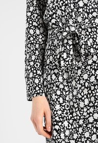 Vestido negro con un estampado floral blanco, con mangas largas y un detalle de cintura atada. La tela parece ligera y texturizada.