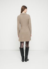 Nő bézs színű bordázott kötött ruhában, fekete harisnyában, amelyen szívminták futnak végig a hátsó részen, és fekete térdig érő csizmában, fehér fal előtt áll.