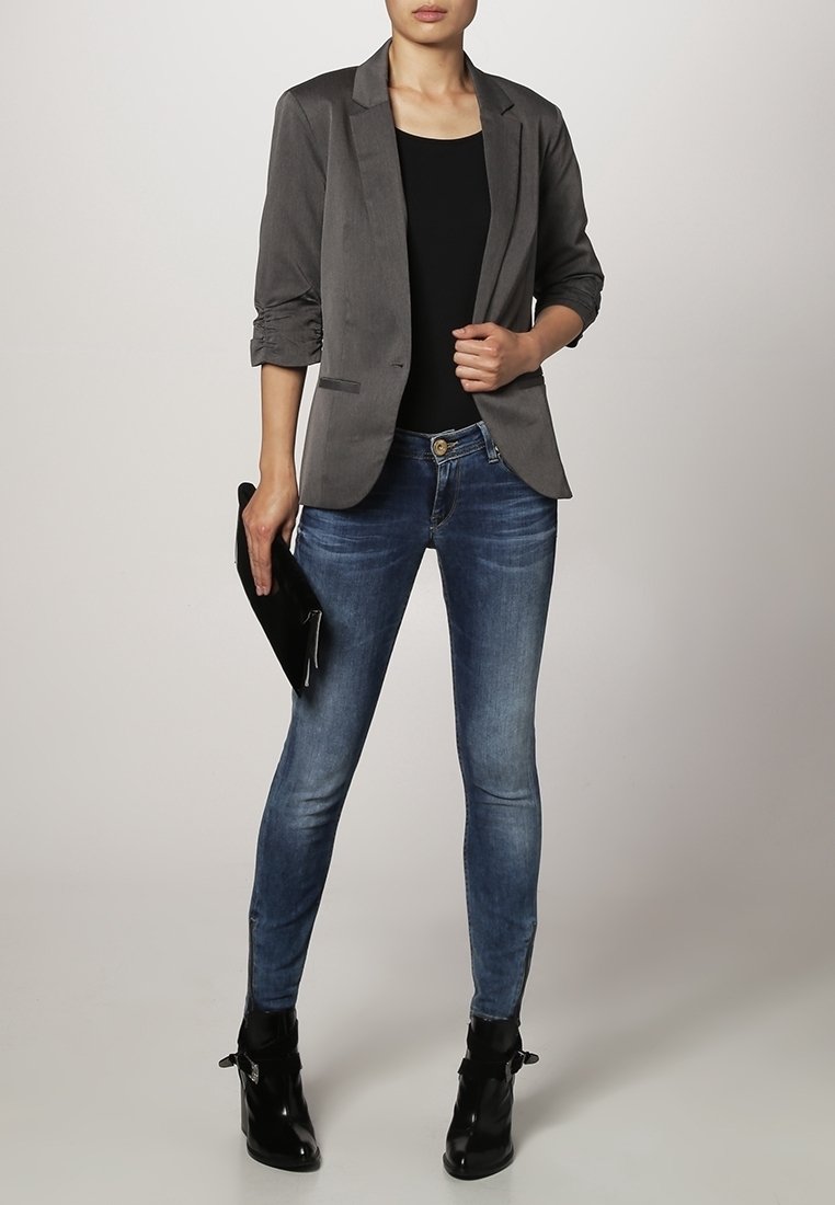 Femme portant un blazer gris, un top noir, un jean skinny bleu et des bottines noires, tenant une pochette noire dans une main.