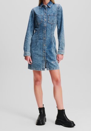 Femme portant une robe en denim bleu ajustée avec des manches longues et des bottines noires épaisses, debout devant un fond uni.