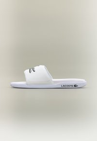 Ciabatte bianche con suola piatta, presentano una fascia testurizzata e un logo Lacoste verde sul lato. Realizzate in materiale sintetico.