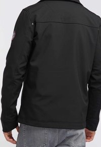 Veste noire à texture lisse avec un col, dotée d'un patch de marque sur l'épaule gauche et de détails de coutures visibles à l'arrière.