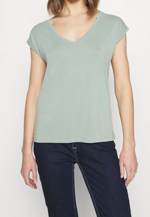 Nainen, joka pukeutunut vaaleanvihreään, lyhythihaiseen V-kaulaiseen paitaan ja tummansinisiin farkkuihin, joissa on valkoiset saumat, kuvattu hartioista lantioihin.