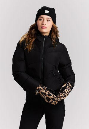 Frau trägt eine schwarze Mütze, eine schwarze Steppjacke und Handschuhe mit Leopardenmuster, steht vor einem einfachen hellen Hintergrund.
