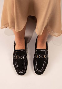 Zapatos de ante negros sin cordones con detalles de tachuelas metálicas y un patrón de diamante en la correa. Textura suave, diseño de punta redonda.