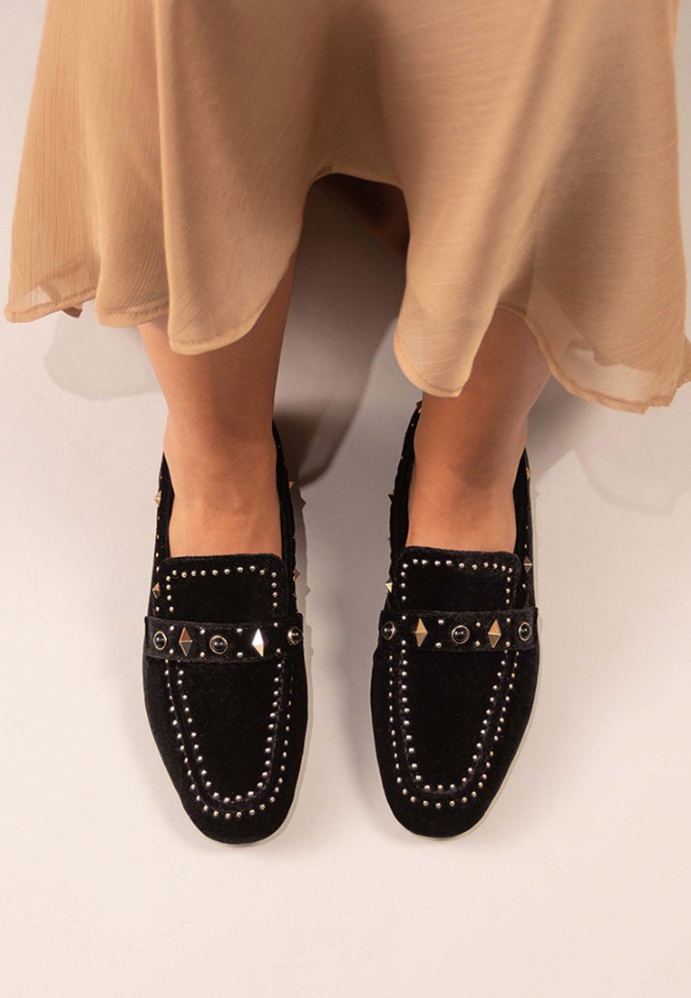 Zapatos de ante negros sin cordones con detalles de tachuelas metálicas y un patrón de diamante en la correa. Textura suave, diseño de punta redonda.
