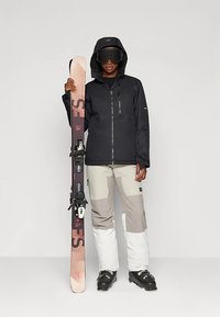 Murtosuojainen lasketteluasu, johon kuuluu musta vedenpitävä takki, harmaat ja valkoiset housut sekä mustat saappaat. Yhdessä kädessä pidetään suksea, jossa on vaaleanpunainen kuvio.