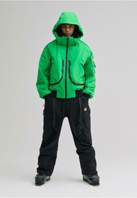 Giacca impermeabile verde brillante con cappuccio, chiusura con zip e molte tasche, abbinata a pantaloni neri e stivali grigi. Texture liscia.