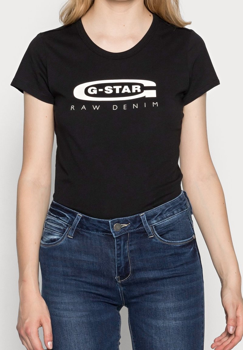 Sort kortærmet t-shirt med hvidt "G-STAR RAW DENIM"-logo, lavet af bomuld, tætsiddende design, kombineret med mørkeblå jeans.