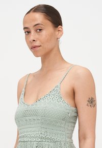 Mentazöld csipke ruha, amely illeszkedő felsőrésszel, pántos vállpántokkal és a szöveten végigfutó bonyolult horgolt mintákkal rendelkezik.