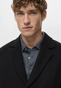 Abrigo de lana negro con solapas sobre una camisa gris. El abrigo tiene una textura suave, mientras que la camisa presenta un diseño con cuello y botones.