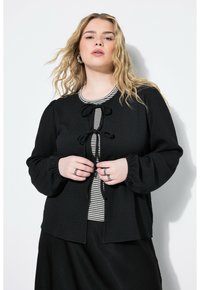 Giacca nera con texture, maniche a sbuffo e chiusura frontale con fiocco. Indossata sopra una camicia a righe, che mette in mostra un design a strati.