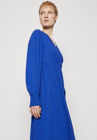 Abito lungo a maniche lunghe di un blu reale con scollatura a V e tessuto strutturato. Presenta polsini arricciati e una vita aderente con una gonna fluida.