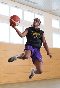 Basketbola spēlētājs melnā bez piedurknēm kreklā un violetās šortos, lēkā, turēdams basketbola bumbu, ar sporta apaviem un bizēm.