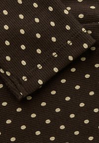 Brunt ribbet stoff med jevnt fordelte kremfargede polkadotter, med synlige sømdetaljer på mansjetten og jevn tekstur over hele.
