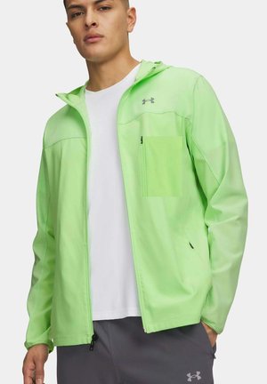 Junger Mann trägt eine hellgrüne Jacke mit Reißverschluss über einem weißen Hemd und dunkelgrauen Hosen, die Hände in den Taschen.