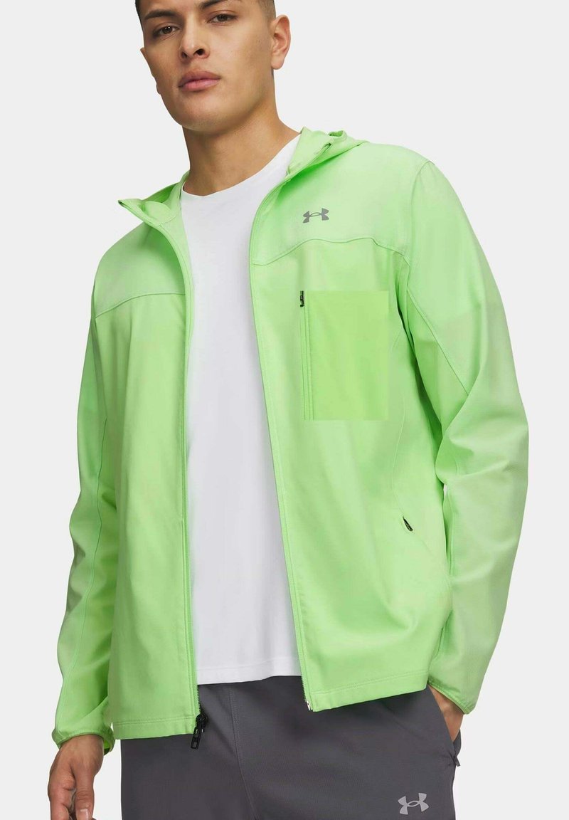 Jonge man draagt een lichtgroene jas met ritssluiting over een wit shirt en donkergrijze broek, met handen in de zakken.