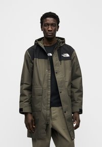 Πράσινο παλτό παρκά ελιάς με μαύρες λεπτομέρειες, ρυθμιζόμενο κουκούλα, εμπρόσθιες τσέπες και κλείσιμο με κουμπιά, κατασκευασμένο από αδιάβροχο υλικό.