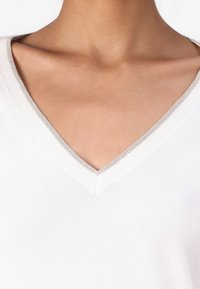Pull blanc en V avec une texture côtelée et un accent argenté au niveau du col, mettant en valeur un tissu lisse et doux.