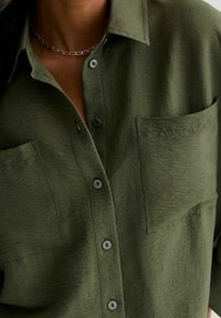 Olivgrön knäppskjorta med två bröstfickor, bärs med ett tunt silversmycke, på en person.