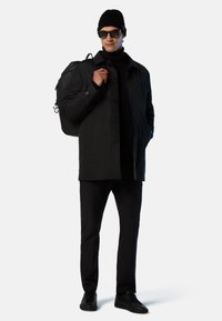 Black waterproof jacket with button closures, black knit turtleneck, black pants, and black sneakers; carrying a black backpack.
