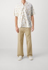 Camicia a maniche corte con colletto di colore bianco con motivo floreale beige; abbinata a pantaloni larghi color khaki chiaro e scarpe da ginnastica bianche.