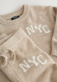 Bežani pleteni pulover i odgovarajuće hlače s bijelim natpisom "NYC". Mekana tekstura, rebrasti rubovi i klasična okrugla izreza.